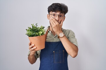 Poster - Arab man with beard holding green plant pot shocked covering mouth with hands for mistake. secret concept.