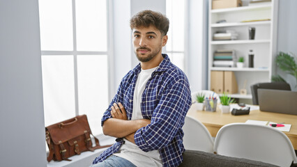 Canvas Print - Confident young arab man exhibits professional success, positively embracing his job as he sits in the office with crossed arms gesture