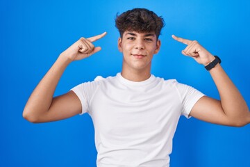 Canvas Print - Hispanic teenager standing over blue background smiling pointing to head with both hands finger, great idea or thought, good memory