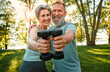 Motivated couple training together. Portrait of happy spouses standing in park and posing with two dumbbells in hands. Cheerful male and female dressed in sport outfit looking at camera and smiling.