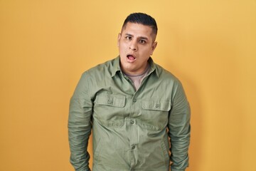 Wall Mural - Hispanic young man standing over yellow background in shock face, looking skeptical and sarcastic, surprised with open mouth