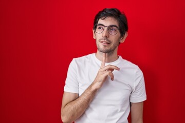 Poster - Young hispanic man standing over red background thinking concentrated about doubt with finger on chin and looking up wondering