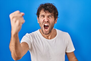 Poster - Hispanic young man standing over blue background angry and mad raising fist frustrated and furious while shouting with anger. rage and aggressive concept.