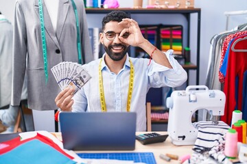 Canvas Print - Hispanic man with beard dressmaker designer holding dollars smiling happy doing ok sign with hand on eye looking through fingers