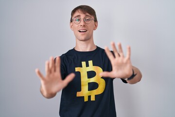 Poster - Caucasian blond man wearing bitcoin t shirt afraid and terrified with fear expression stop gesture with hands, shouting in shock. panic concept.