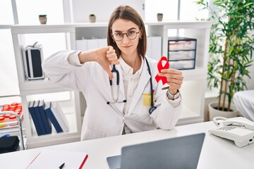 Poster - Young caucasian doctor woman holding support red ribbon with angry face, negative sign showing dislike with thumbs down, rejection concept