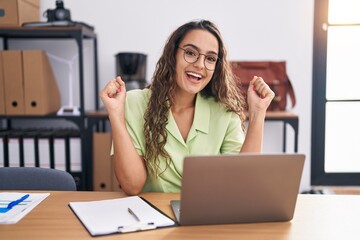 Sticker - Young hispanic woman working at the office wearing glasses celebrating surprised and amazed for success with arms raised and open eyes. winner concept.