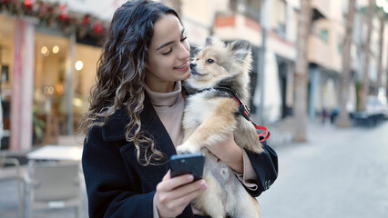 Wall Mural - Young hispanic woman with dog smiling confident using smartphone at street