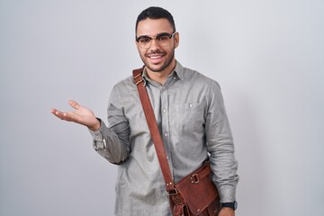 Wall Mural - Young hispanic man wearing suitcase smiling cheerful presenting and pointing with palm of hand looking at the camera.