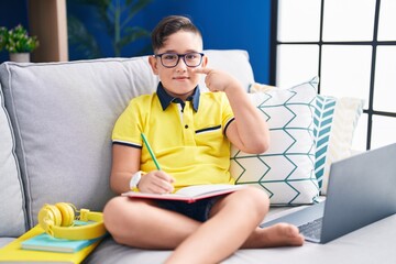 Poster - Young hispanic kid doing homework sitting on the sofa pointing with hand finger to face and nose, smiling cheerful. beauty concept