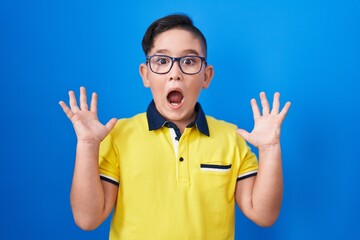 Poster - Young hispanic kid standing over blue background celebrating victory with happy smile and winner expression with raised hands