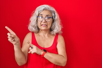 Canvas Print - Middle age woman with grey hair standing over red background pointing aside worried and nervous with both hands, concerned and surprised expression