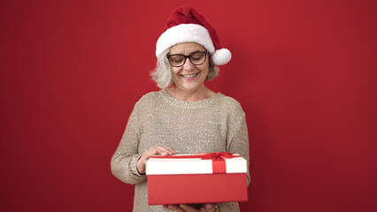 Sticker - Middle age woman with grey hair wearing christmas hat unpacking gift over isolated red background