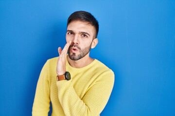 Poster - Hispanic man standing over blue background hand on mouth telling secret rumor, whispering malicious talk conversation