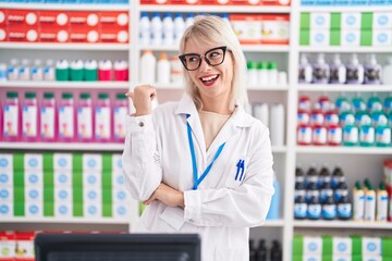 Poster - Young caucasian woman working at pharmacy drugstore smiling with happy face looking and pointing to the side with thumb up.