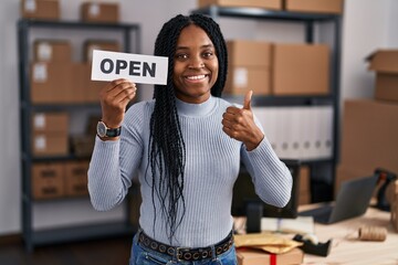 Wall Mural - African american woman working at small business ecommerce holding open banner smiling happy and positive, thumb up doing excellent and approval sign