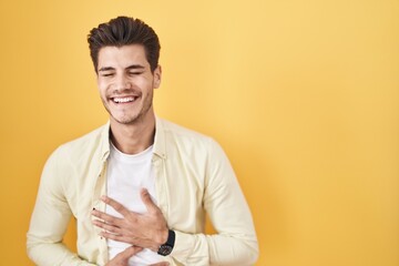 Poster - Young hispanic man standing over yellow background smiling and laughing hard out loud because funny crazy joke with hands on body.