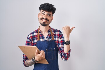 Sticker - Young hispanic man with beard wearing waiter apron holding clipboard smiling with happy face looking and pointing to the side with thumb up.