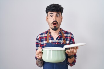 Poster - Young hispanic man with beard wearing apron holding cooking pot afraid and shocked with surprise and amazed expression, fear and excited face.