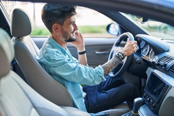 Wall Mural - Young hispanic man talking on smartphone sitting on car at street