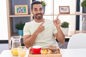 Canvas Print - Hispanic man with beard eating breakfast smiling and looking at the camera pointing with two hands and fingers to the side.