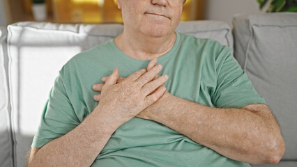 Wall Mural - Middle age grey-haired man sitting on sofa with hands on heart at home