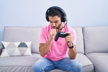 Wall Mural - Hispanic young man playing video game holding controller sitting on the sofa feeling unwell and coughing as symptom for cold or bronchitis. health care concept.