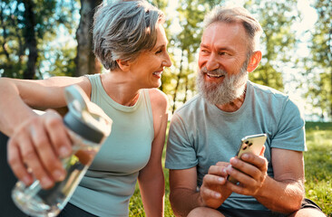 Wall Mural - Carefree and active retirement life. Happy aged couple resting after training on nature and smiling while looking at each other. Joyful woman holding water bottle while man showing pics on smartphone.