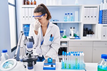 Wall Mural - Young hispanic woman working at scientist laboratory tired rubbing nose and eyes feeling fatigue and headache. stress and frustration concept.