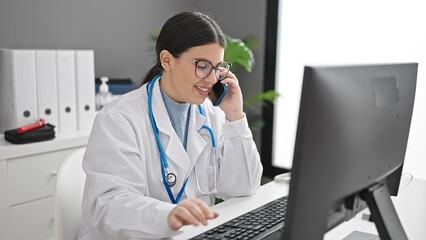 Sticker - Young hispanic woman doctor using computer speaking on smartphone at clinic