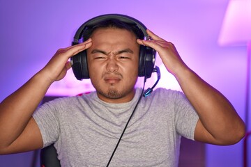 Poster - Chinese young man playing video games wearing headphones with hand on head, headache because stress. suffering migraine.