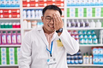 Sticker - Chinese young man working at pharmacy drugstore covering one eye with hand, confident smile on face and surprise emotion.