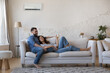 Happy millennial husband and wife resting on couch at home, relaxing under cooling air from conditioner, holding remote control for AC, using domestic appliance for comfort
