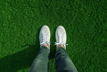 Close-up Of Female Legs In White Sneakers On Green Grass Background