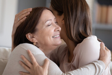  Adult daughter embraces middle-aged 50s mother different generation age women seated on sofa at home close up. Enjoy meeting, time together, showing each other love care and unconditional love concept