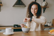 © ViDi Studio - Young smiling woman wears casual clothes sweater eat breakfast muesli cereals with milk fruit in bowl look at smart watch sit at table in light kitchen at home alone. Lifestyle cooking food concept.