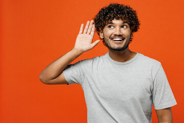 Wall Mural - Young curious nosy cheerful fun happy Indian man he wears t-shirt casual clothes try to hear you overhear listening intently isolated on orange red color background studio portrait. Lifestyle concept.