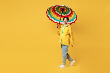 Wall Mural - Full body side view young woman wear waterproof raincoat outerwear red hat hold umbrella walk go isolated on plain yellow background studio portrait. Outdoor lifestyle wet fall weather season concept