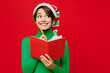 © ViDi Studio - Young woman wear green turtleneck Santa hat posing hold in hand diary notebook write down dreams look aside isolated on plain red background. Happy New Year 2024 celebration Christmas holiday concept.