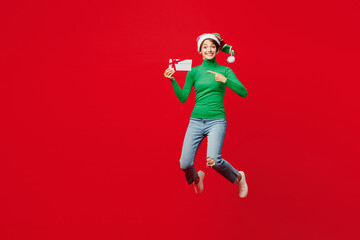 Wall Mural - Full body young woman wear green turtleneck Santa hat posing jump high hold store gift coupon voucher card isolated on plain red background. Happy New Year 2024 celebration Christmas holiday concept.