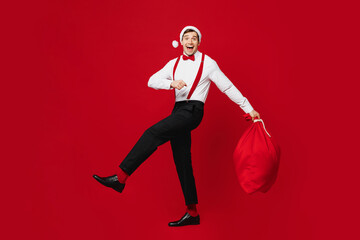 Wall Mural - Full body side view merry excited young man wear white shirt Santa hat posing hold bag with present gifts isolated on plain red background studio. Happy New Year Christmas celebration holiday concept.