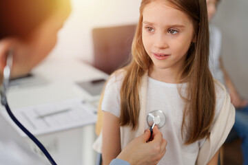 Wall Mural - Doctor and kid patient are in the clinic. Physician in white coat examining a serious young girl with a stethoscope, close up. Medicine, therapy concept