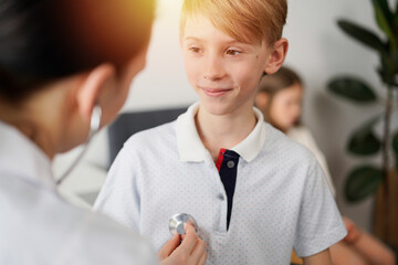 Wall Mural - Doctor and kid patient are in the clinic. Physician in white coat examining a smiling young boy with a stethoscope, close up. Medicine, therapy concept