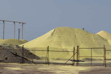 Mounds of yellow sulfur powder behind a metal fence. The pile is used for oil refinement as a waste product of the fossil fuel industry. The stockpile of the chemical sulfate is for bulk storage.