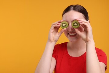 Poster - Smiling woman covering eyes with halves of kiwi on orange background, space for text