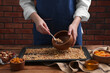 Woman putting granola from baking tray into bowl at wooden table, closeup