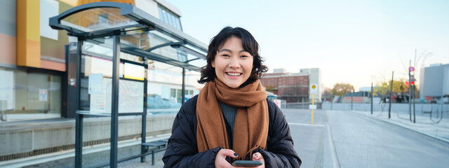 Wall Mural - Portrait of Korean woman in winter jacket, standing with smartphone, waiting for bus on stop, looking at mobile phone app checking public transport application