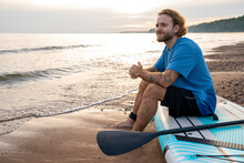 Smiling Man With Tattoo Sitting On Paddleboard At Beach