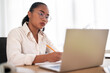 © Studio Marmellata - Focused young female executive in white shirt and eyeglasses working on project using laptop while taking notes in notebook at workplace during workday