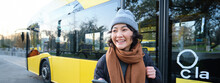Portrait Of Girl Standing Near Bus On A Stop, Waiting For Her Public Transport, Schecks Schedule On Smartphone Application, Holds Mobile Phone, Wears Warm Clothes